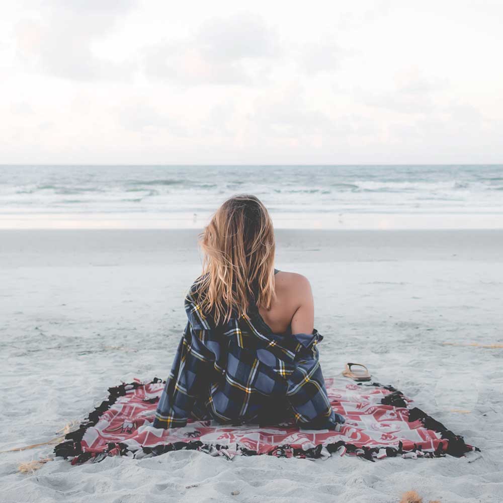 Woman in flannel wrapped in blanket sitting on patterned beach towel watching ocean waves