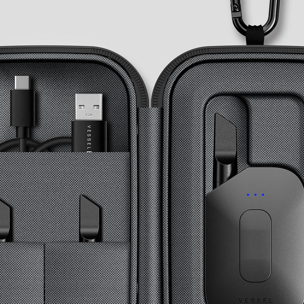 Open charcoal grey and black Vessel Custom Hub case with carabiner displaying Compass Rise, USB-C charging cable, and accessories