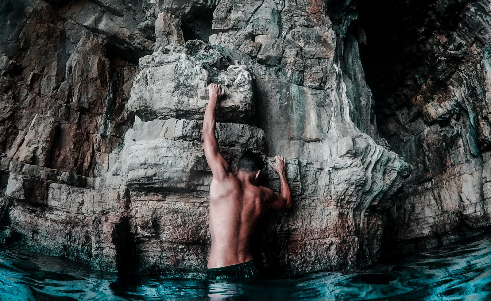 Person climbing a rocky sea cliff from the water, gripping stone ledges inside a coastal cave