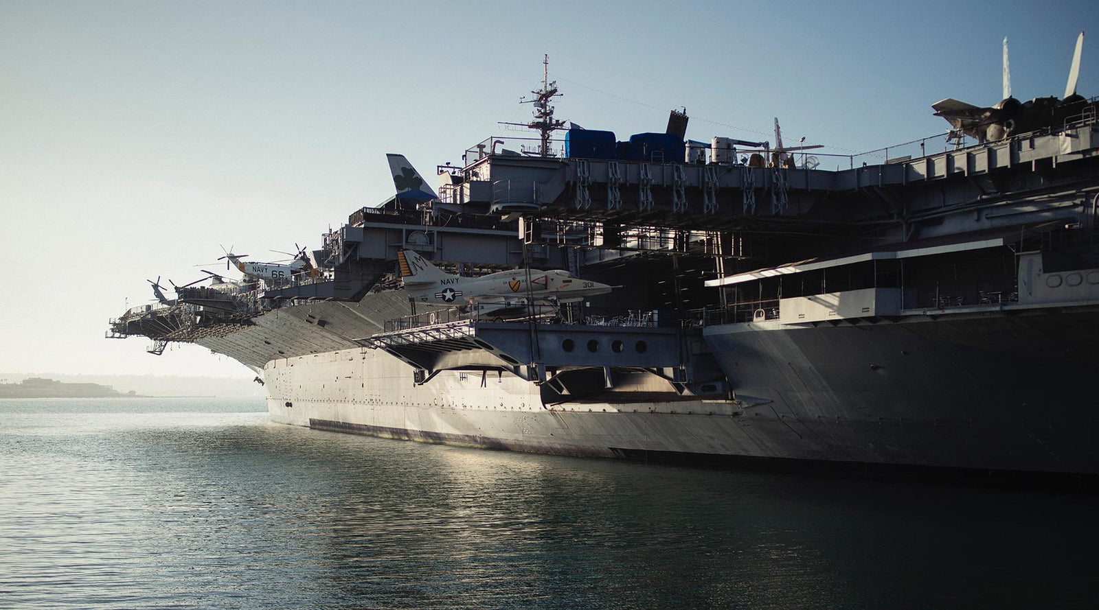 Aircraft carrier docked in calm harbor waters with fighter jets visible on deck